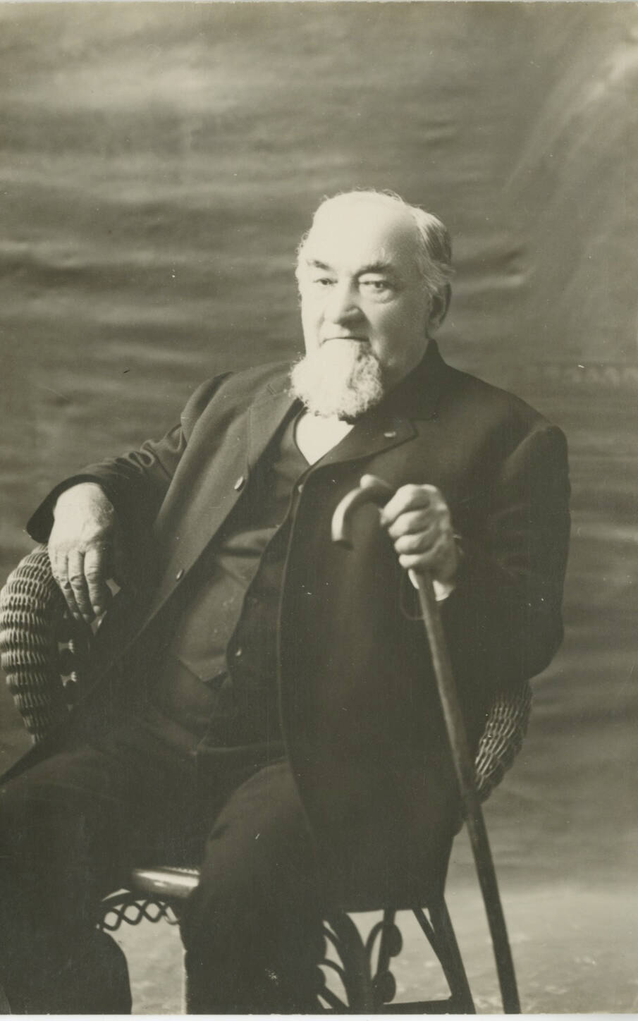 Historic photographic studio portrait of an older man, seated in a chair, holding a cane with a beard.